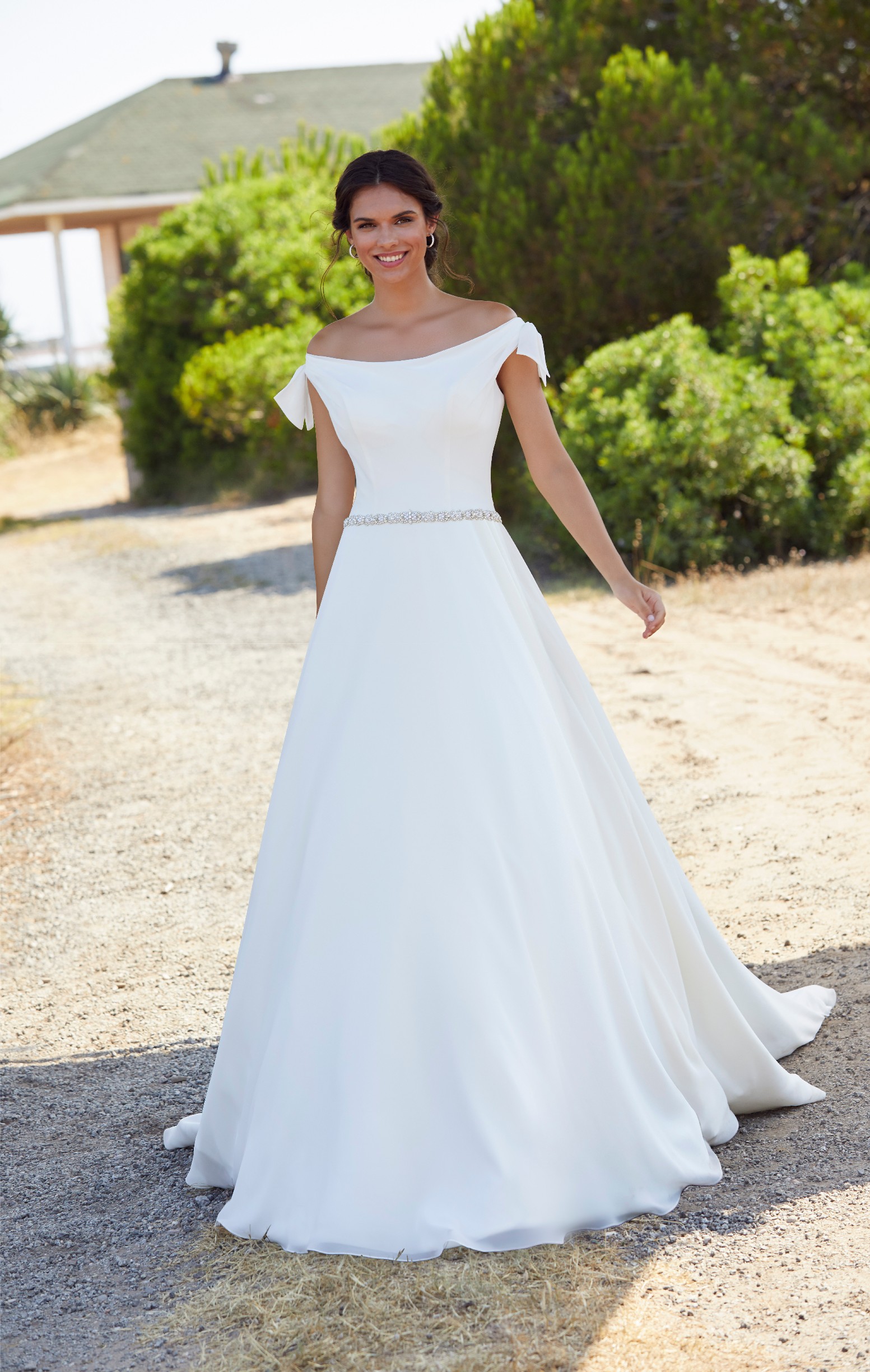 Woman stood in villa garden on sunny day wearing off the shoulder princess style ballgown wedding dress with embellished waist belt detail