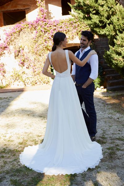 Model stood with a groom in a courtyard wearing Ronald Joyce 18454, a plain wedding dress with a pearl beaded halter neckline and waistband that’s perfect for small weddings