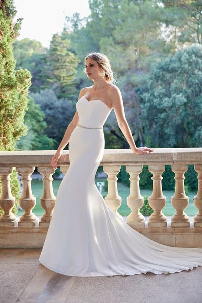 Model stood on a stately home terrace in Ronald Joyce style 18501, a plain fit and flare wedding gown with a strapless sweetheart neckline, sparkle belt and integrated train.