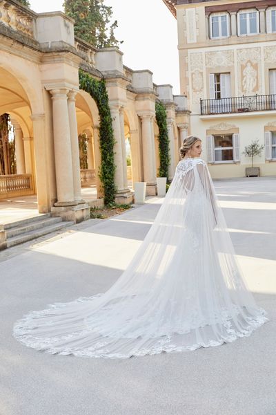 Back of a model stood in a grand Italian courtyard in Ronald Joyce 18503, a vintage inspired lace wedding dress with a fit and flare skirt, v-shaped open back and cathedral-length tulle cape