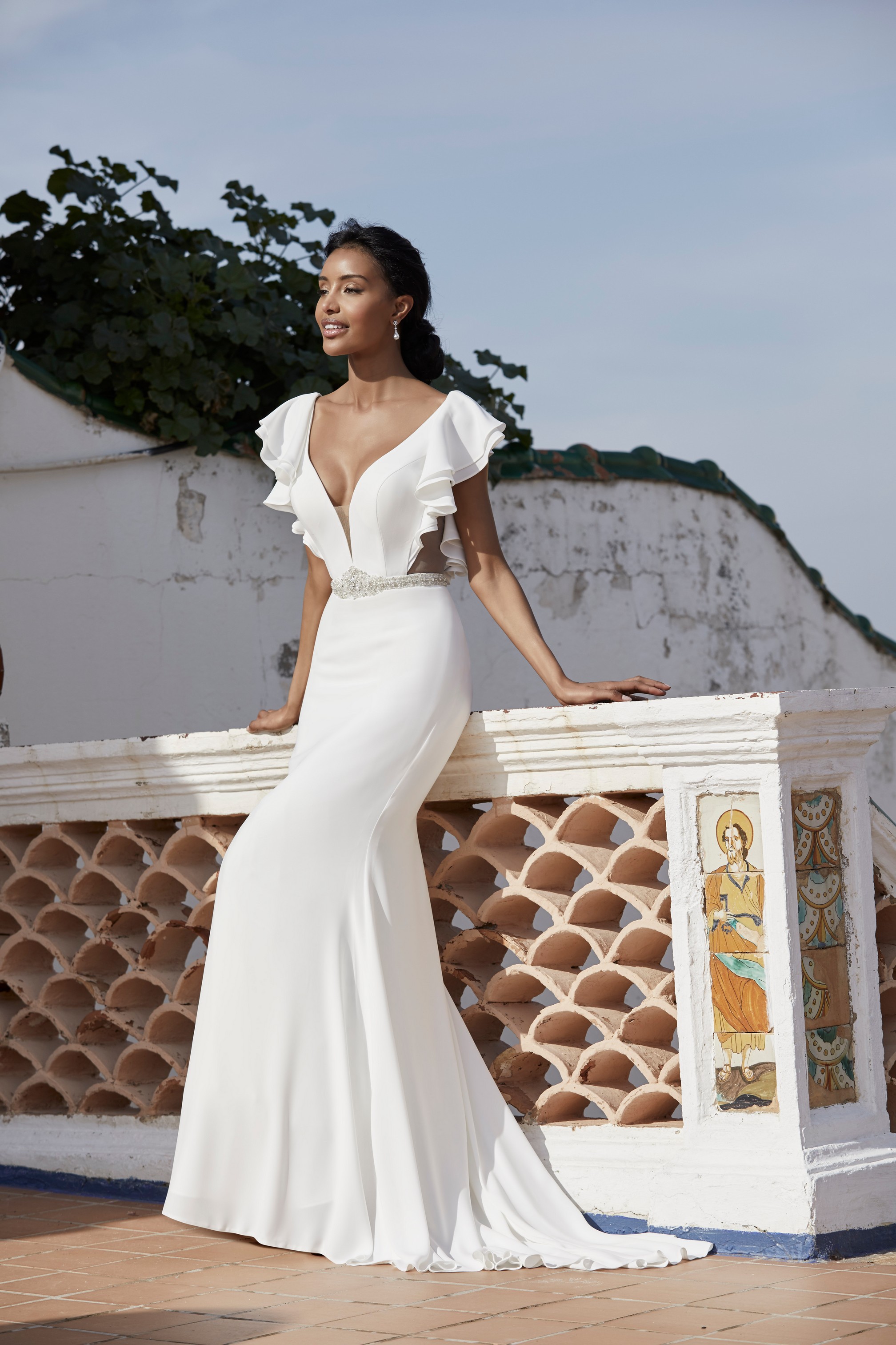 A black model stood on a Spanish terrace in Victoria Jane style 18553, a plain crepe destination wedding gown with a plunging illusion neckline and ruffle cap sleeves 