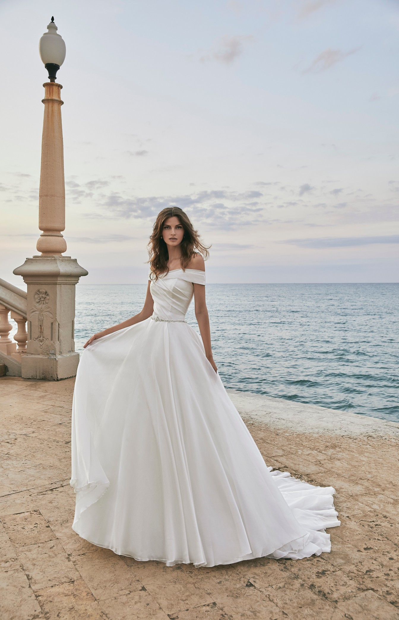 Brunette model stood near a light post by the sea in Victoria Jane 18619, a ballgown wedding dress with a bateau off-the-shoulder neckline and a delicate sparkly diamante belt