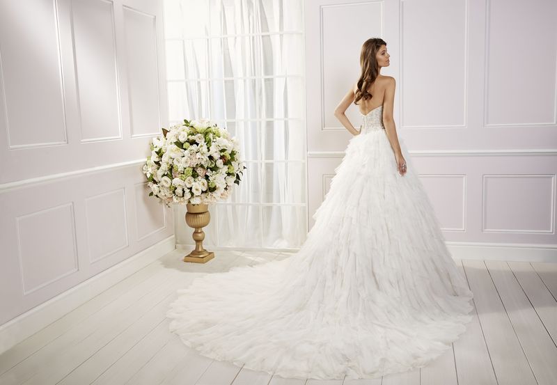 Back image of brunette woman standing in white panelled room in front of floral bouquet display wearing strapless beaded bodice ballgown wedding dress