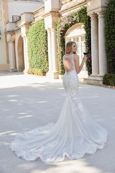 Lady standing outside villa wearing fitted wedding dress with backless detail and lace appliques holding a glass of red wine