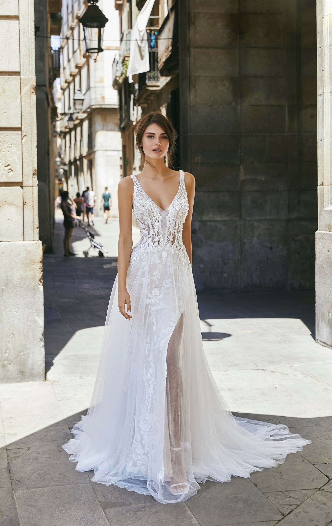 Brunette model stood in a sunny Spanish street in Ronald Joyce 69701, one of our sparkly A-line wedding dresses with a delicately beaded illusion bodice, floral lace appliqués, straps and a glitter tulle skirt with open leg detail.