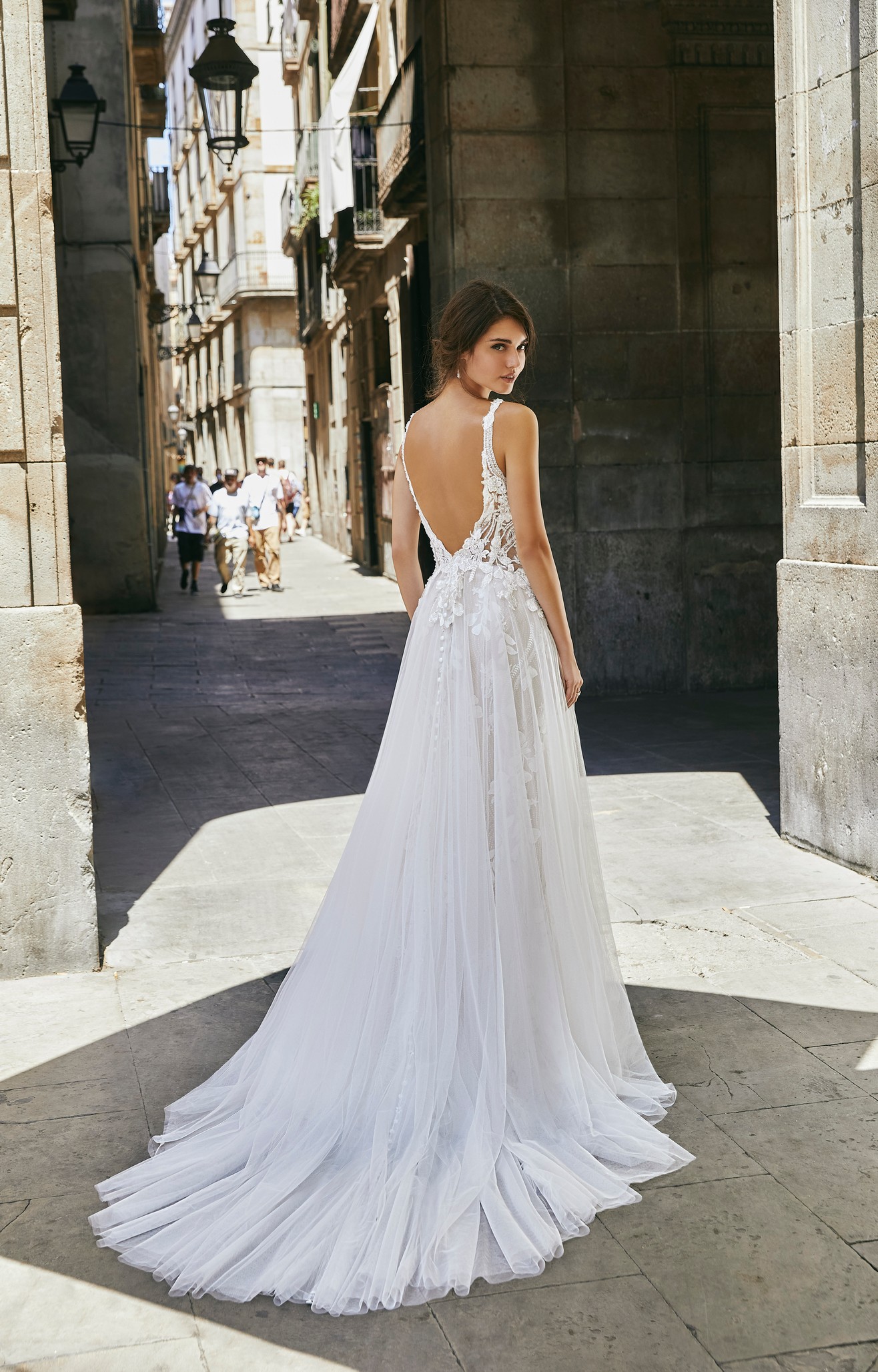 Back of a brunette model stood in a sunny Spanish street in Ronald Joyce 69701, one of our sparkly wedding dresses with an open back, illusion bodice, floral lace appliqués, straps and a glitter tulle skirt with open leg detail.