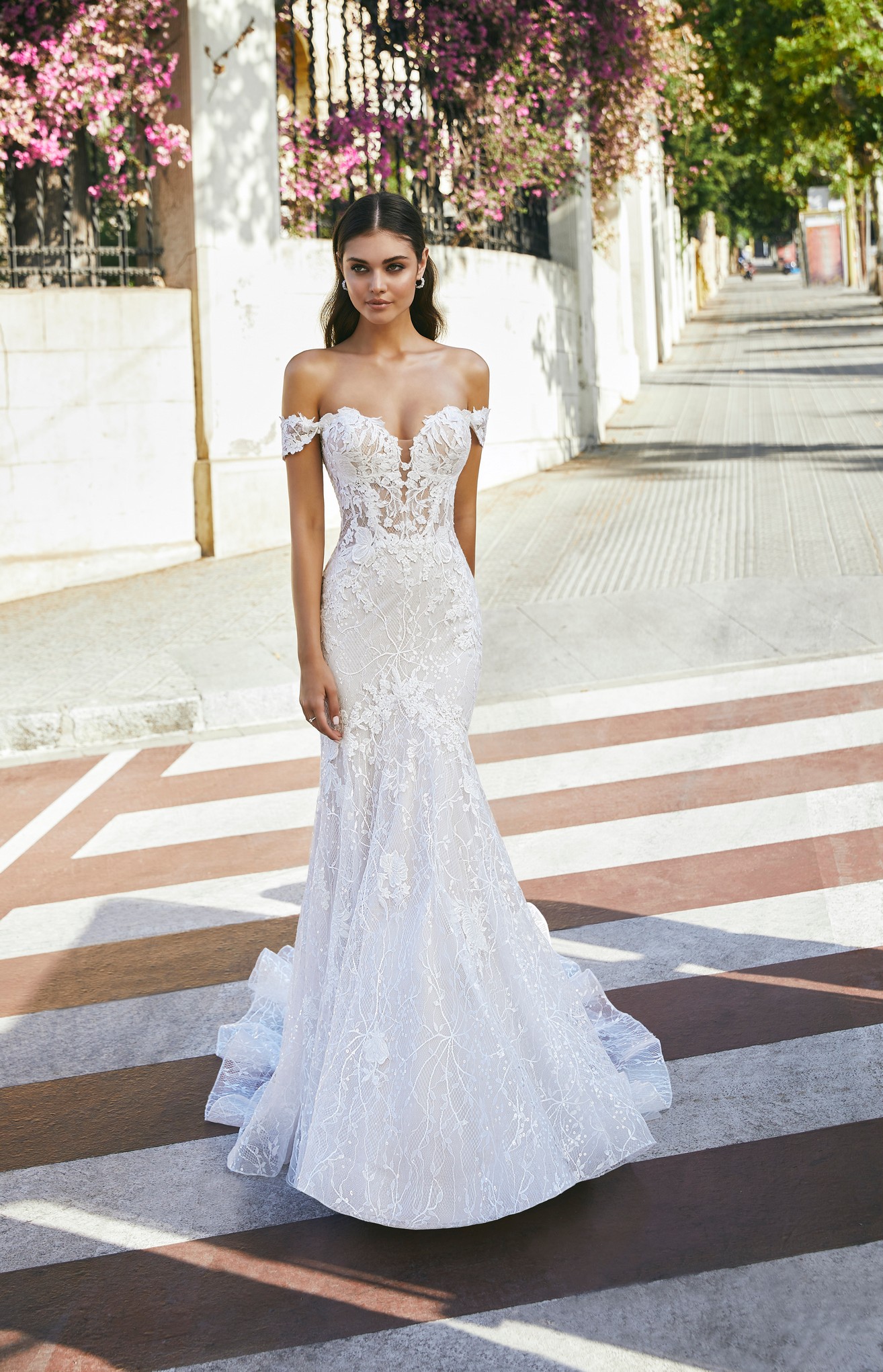 Model stood in a street wearing Ronald Joyce style 69707, a strapless lace fit and flare wedding dress with a plunging sweetheart neckline, illusion bodice and detachable off-the-shoulder short sleeves 