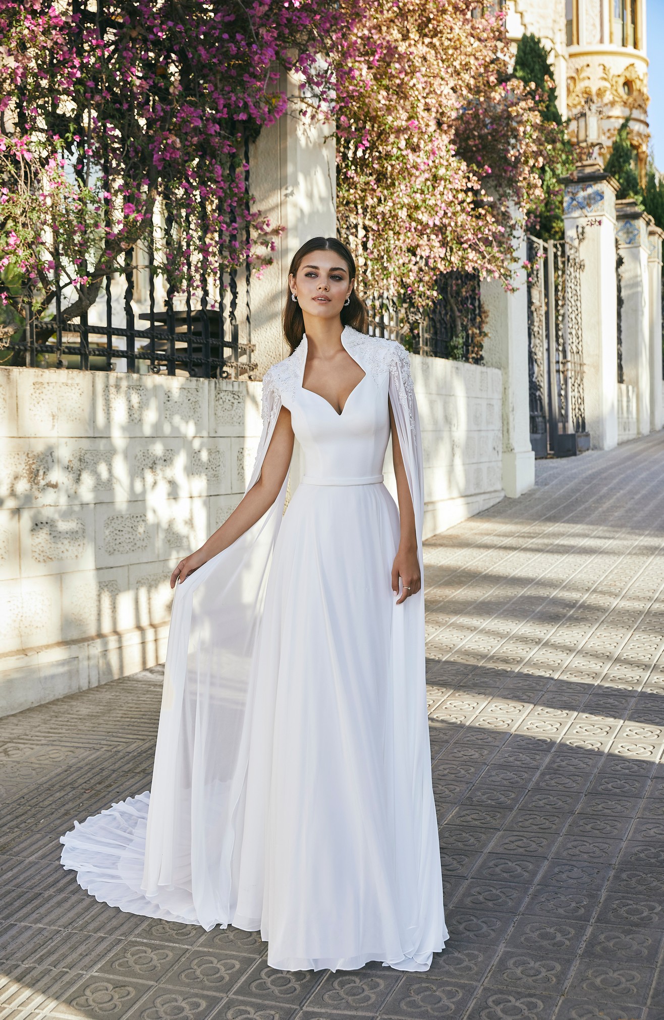 Model stood on a sunny floral Italian path in Ronald Joyce style 69708, an A-line wedding dress with a beaded Queen Anne collar, delicate plain belt and attached chiffon cape complete with beaded shoulder appliques