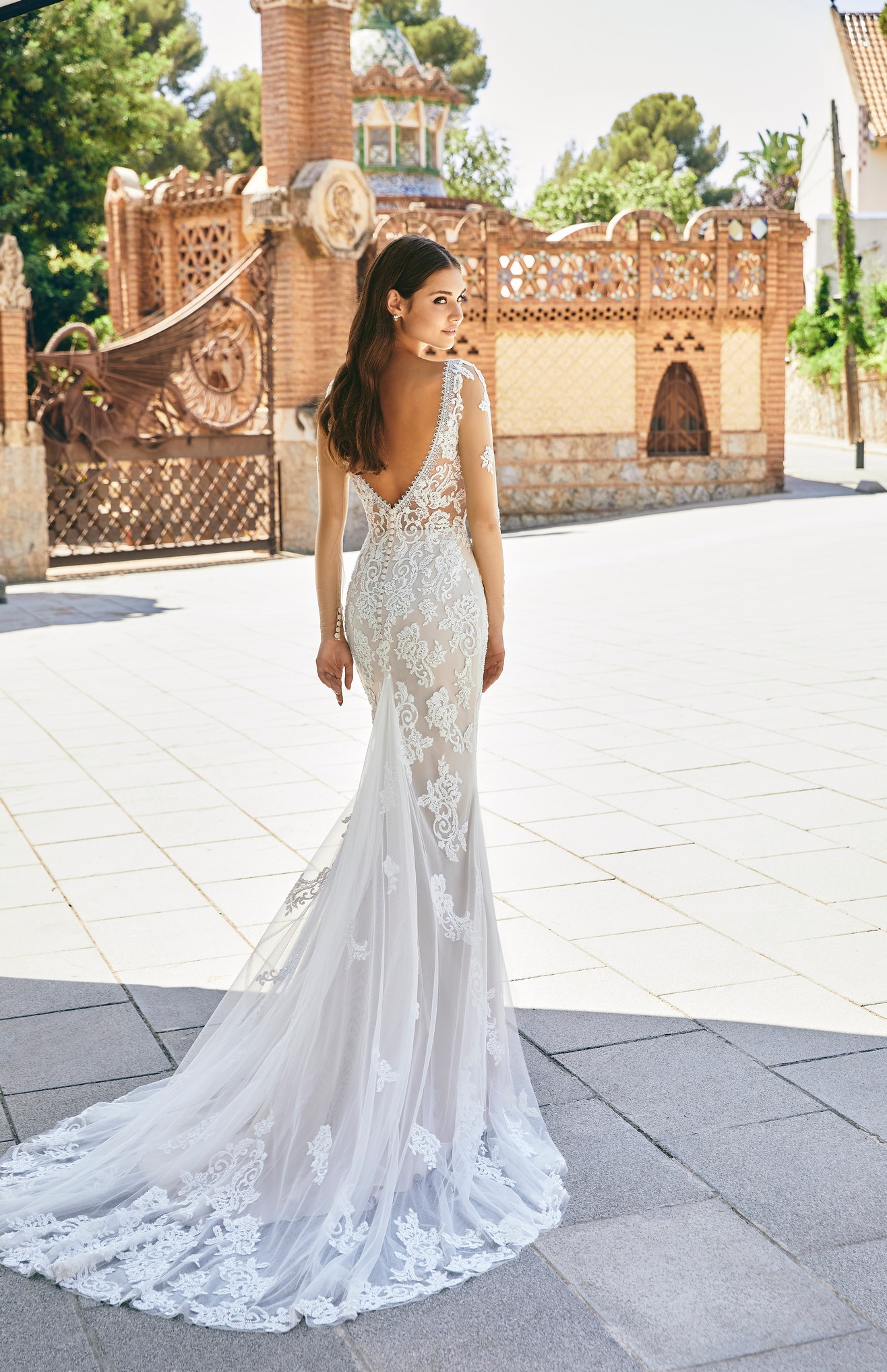 Back of a brunette model stood in a sunny street in Ronald Joyce 69716, a wedding dress with long sheer sleeves, a plunging v-shaped back, illusion bodice and bustle made from tulle and lace applique bridal fabrics