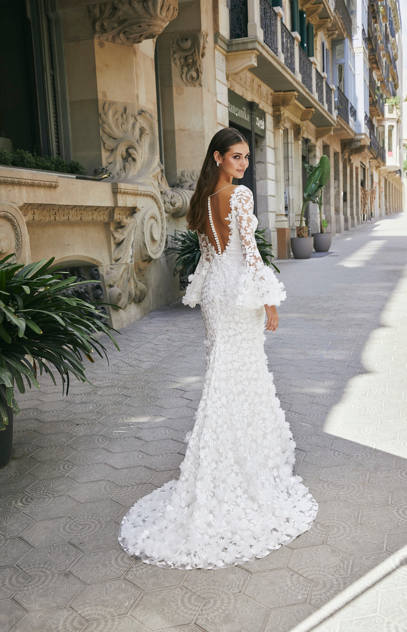 Back of a brunette model stood in an empty sunny street in Ronald Joyce 69725, a boho wedding dress style with 3D floral detail, long fluted sleeves and a buttoned necklace back and puddle train.
