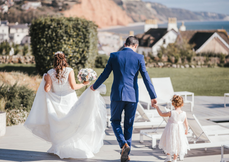 Real bride Sophie walking in Ronald Joyce Robyn wedding dress (style 68060) with her husband Jack and an infant bridesmaid.  Rooftops, a cliff edge and the sea are in the background