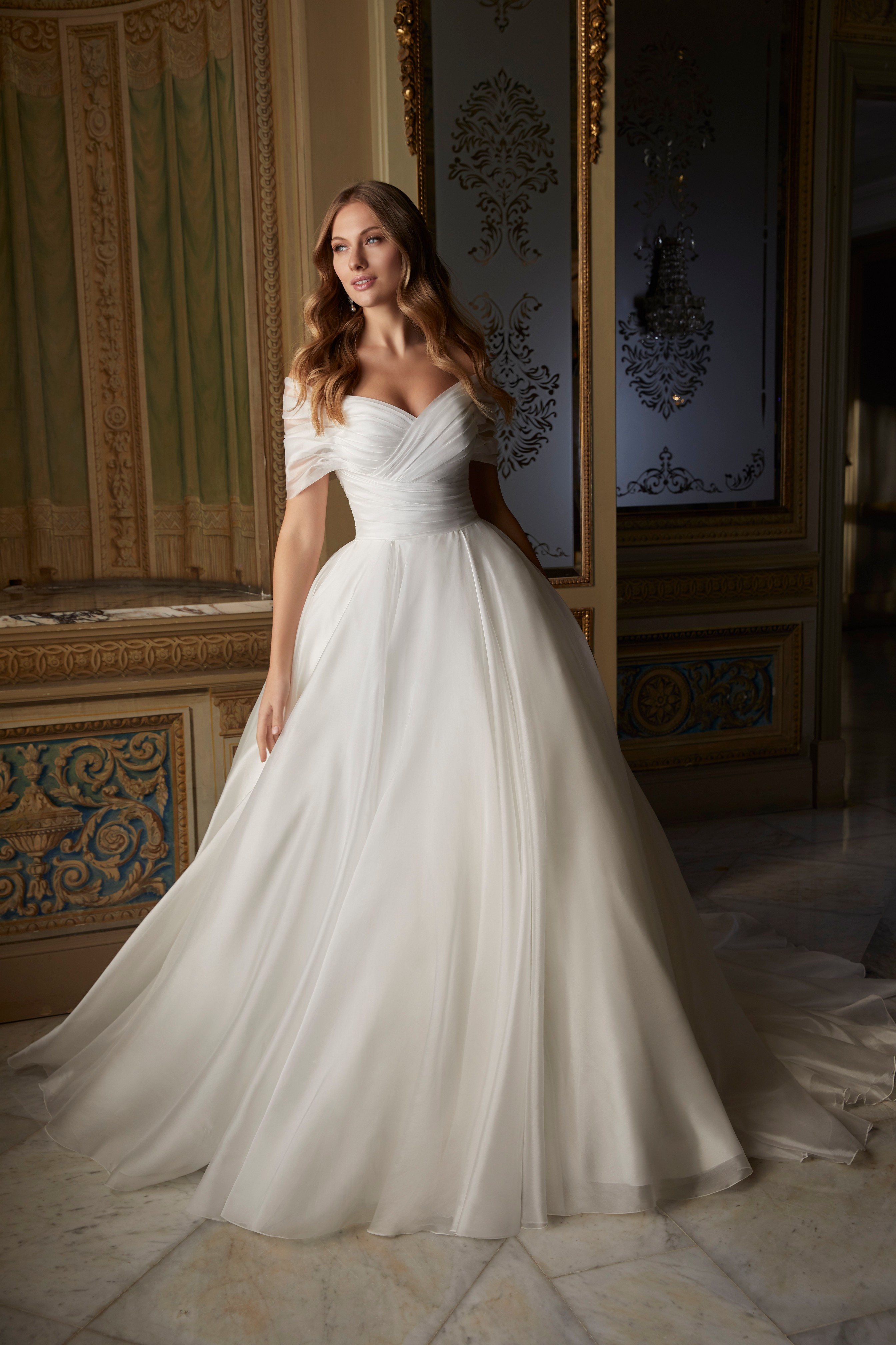 Model stood in a grand building wearing Ronald Joyce style 69551, an elegant plain ballgown wedding dress with off the shoulder sleeves 