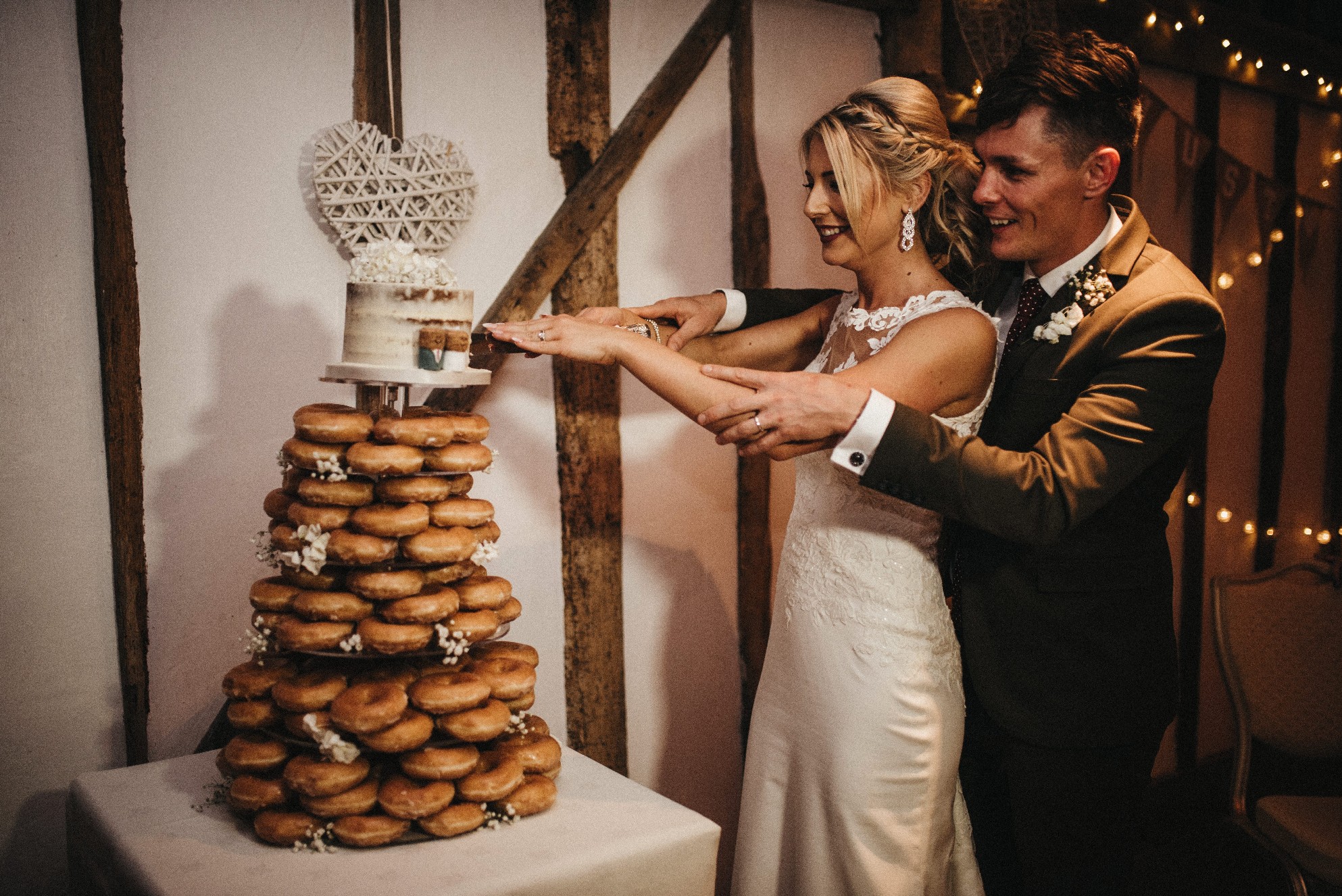 A young bride and groom smiling as they cut a small wedding cake above a tier of doughnuts.jpg