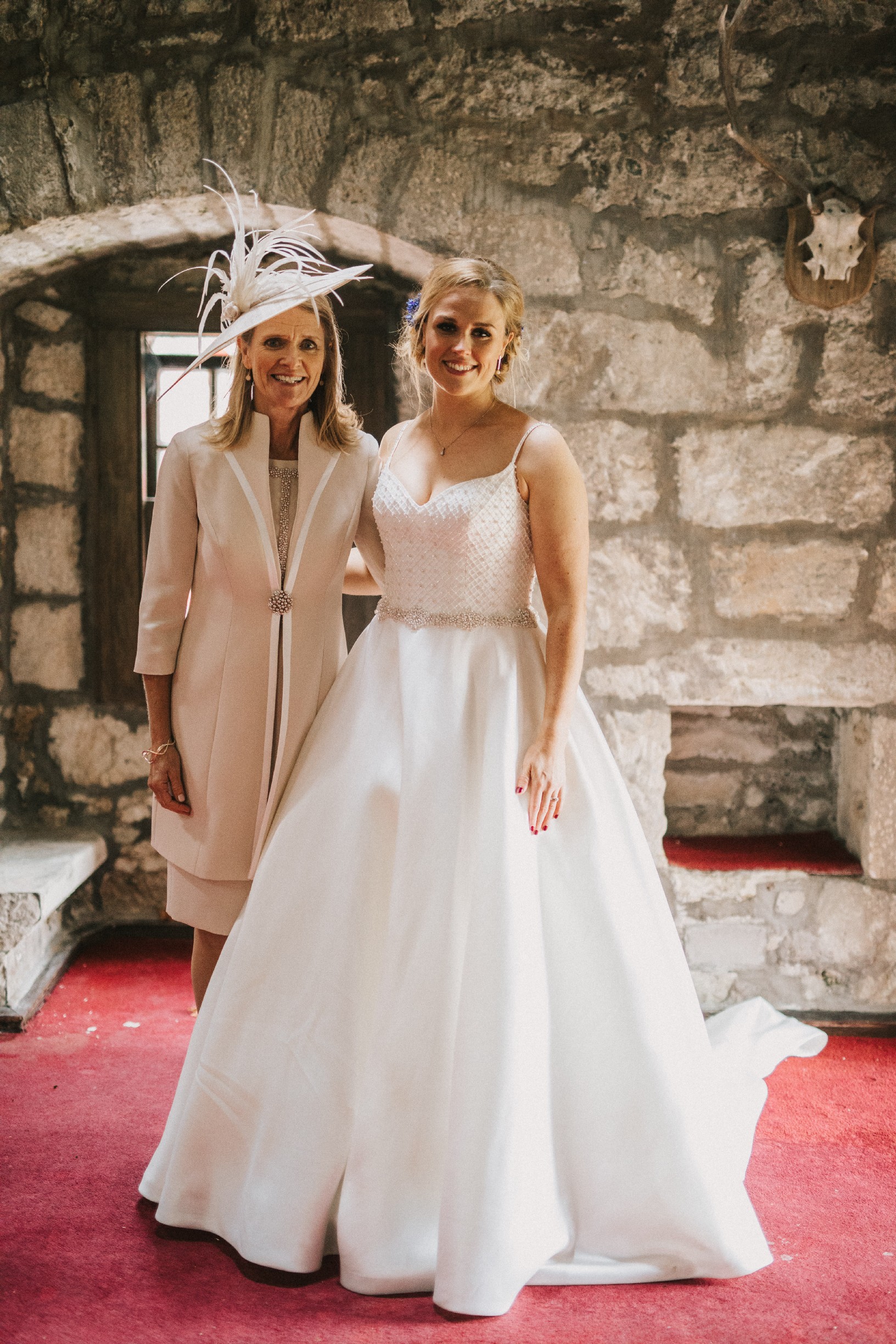 A photo of a bride stood next to her mum inside a castle room wearing an ivory strappy ballgown wedding dress with a sparkle belt