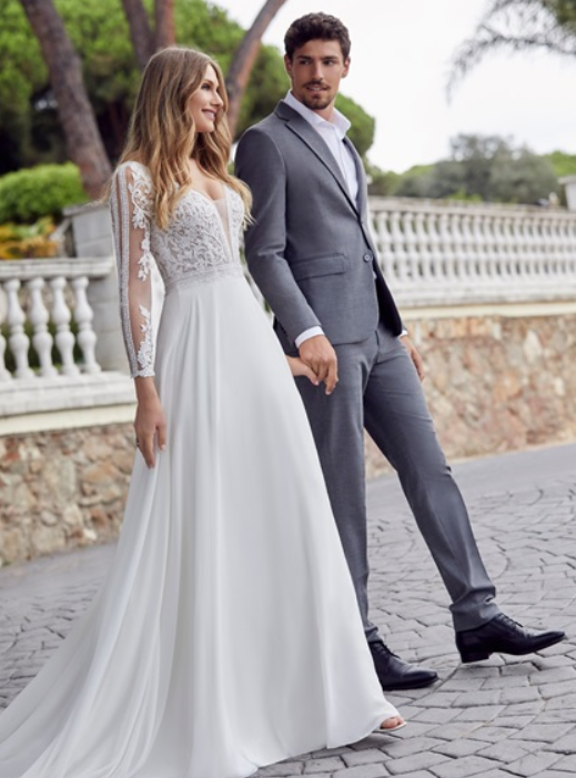 Model stood next to a groom in Ronald Joyce style 69512, an elegant A-line wedding dress with long sheer and lace applique sleeves and a plunging beaded bodice