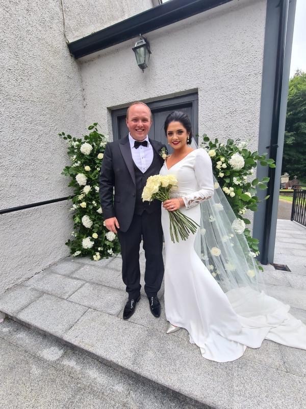 Real bride Emma stood with her husband by a grey building and cream floral bushes. She wears Ronald Joyce 18202, a sheath wedding dress with lace cuff long sleeves, a v-neckline and long train