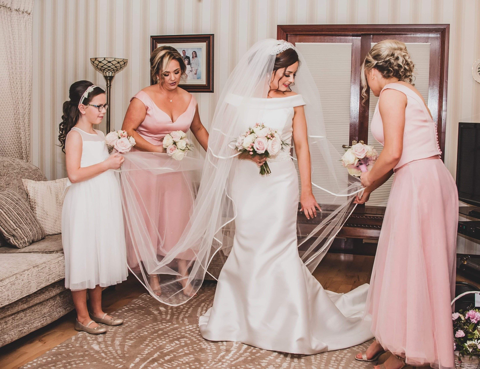 Real bride Sinead stood in a living room with two bridesmaids in pink dresses and a flower girl. Sinead wears an ivory Ronald Joyce 69153 wedding dress and a sweeping tulle veil.