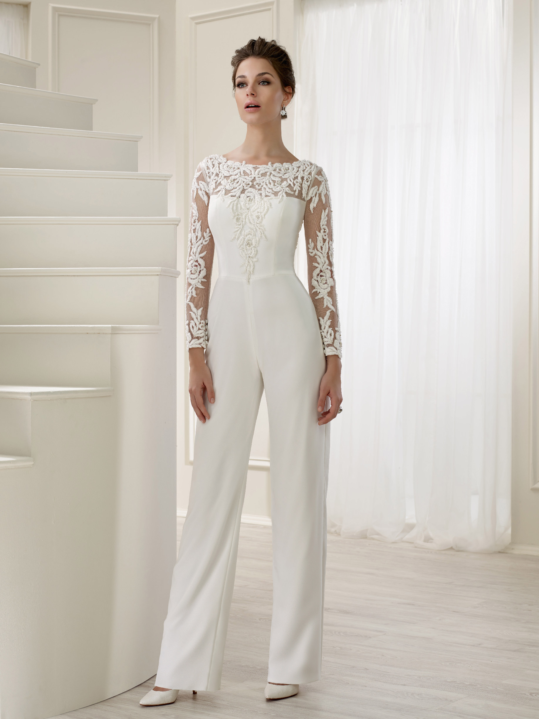 Brunette model stood in a cream room in Ronald Joyce 18161, a plain ivory bridal jumpsuit with sheer lace sleeves and a matching lace illusion neckline – perfect for older brides