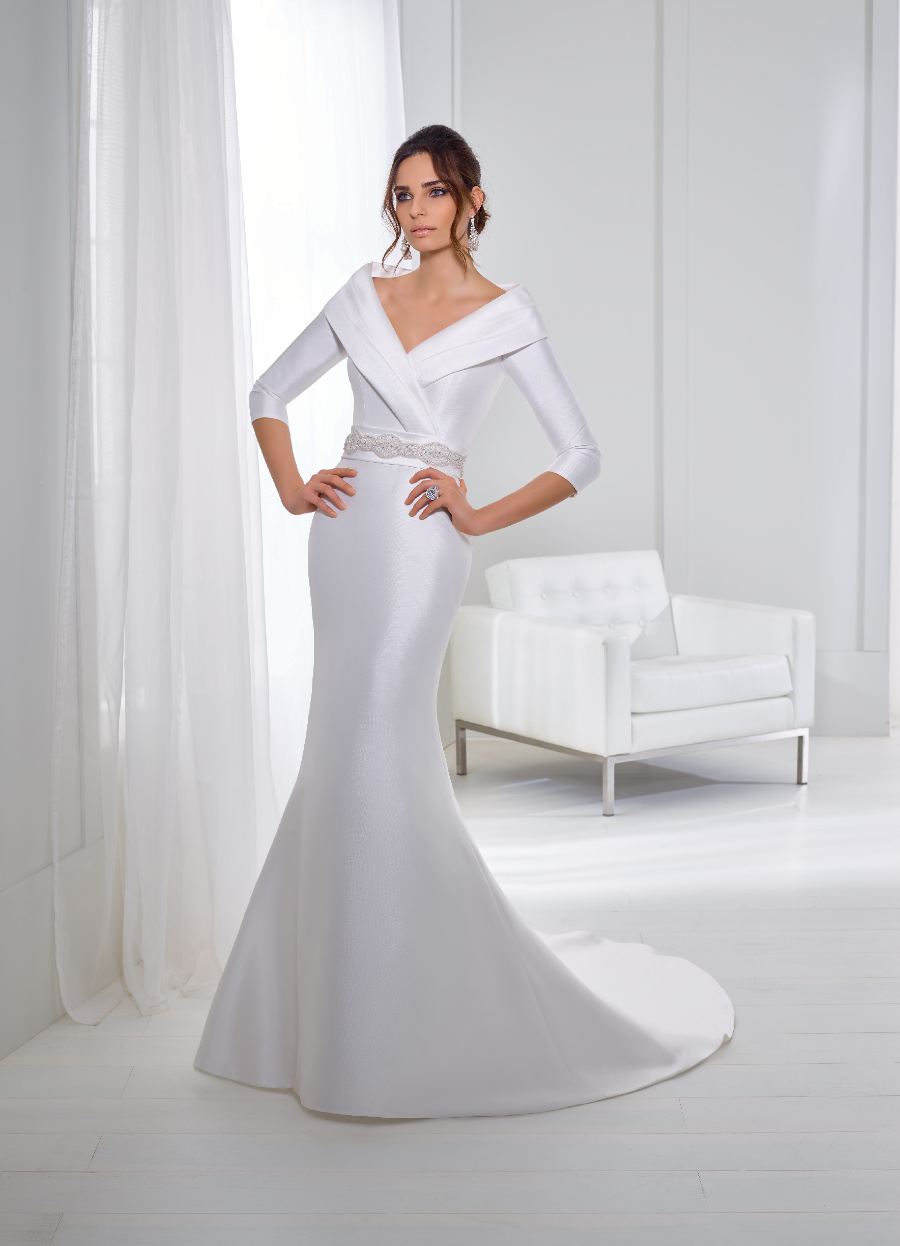 Brunette model stood in a light grey room in Ronald Joyce 18267, a white fishtail Mikado wedding dress with an off the shoulder lapel neckline, ¾ sleeves and sparkle belt 