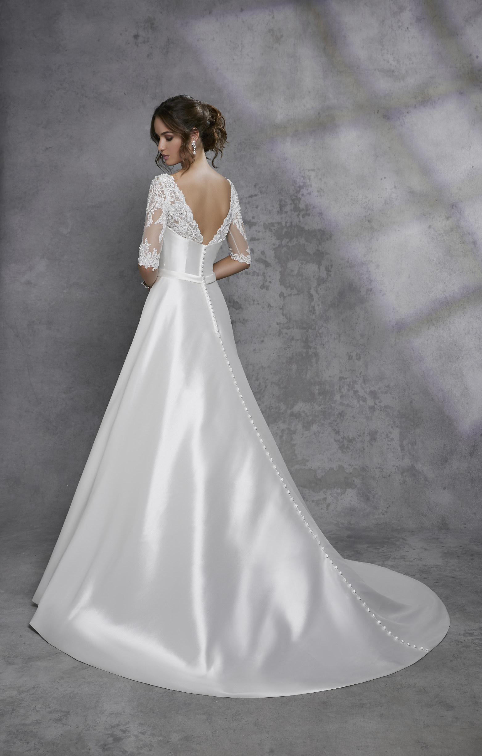 Back image of a woman standing in a photography studio with grey backdrop wearing mikado ballgown wedding dress with low back and lace sleeves
