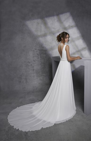 Lady standing in photo studio against a grey backdrop wearing a-line bridal gown with v-shaped back and embellished belt on waistline