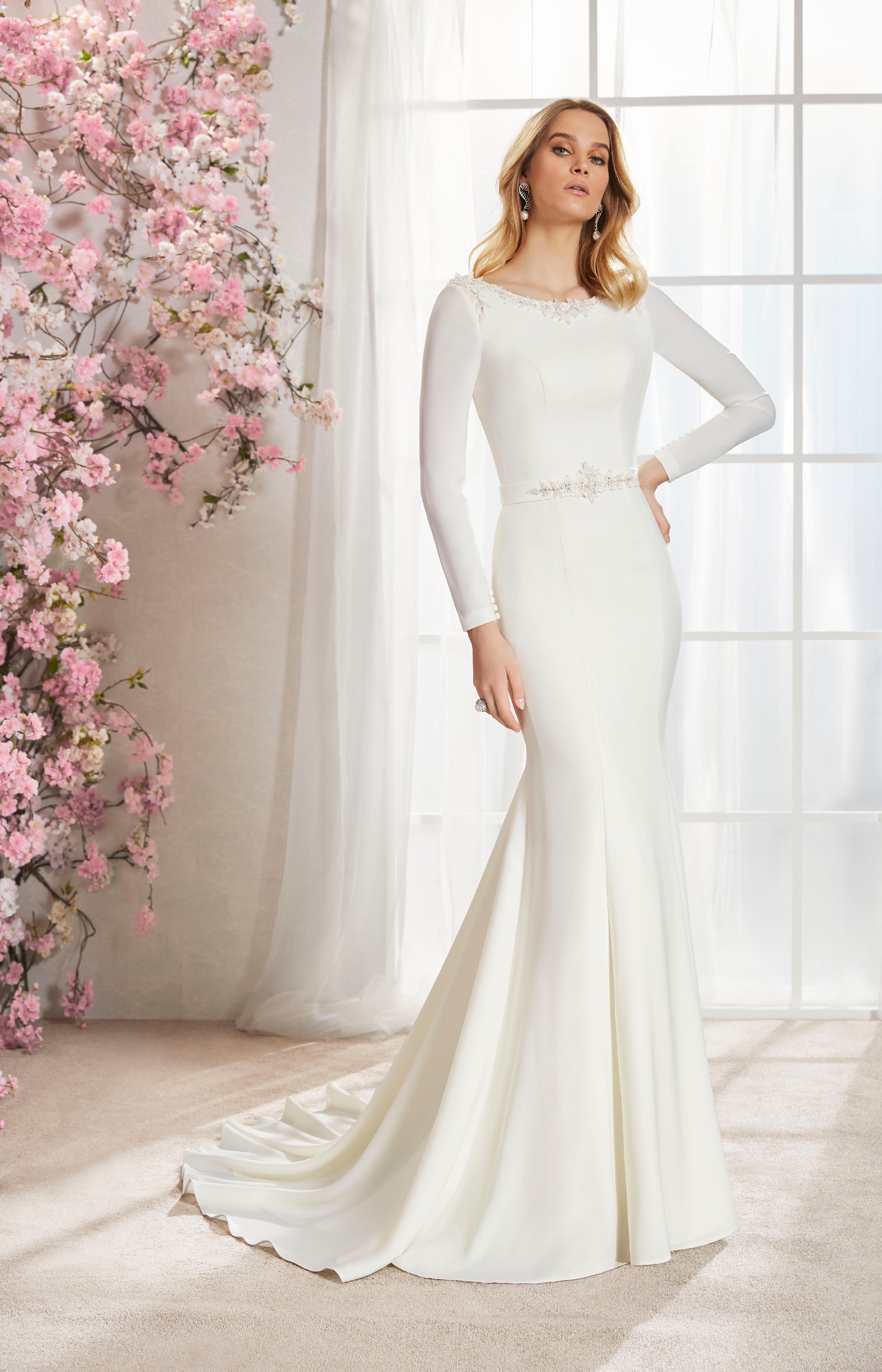 Blonde model stood in a cream room next to pink blossom in Ronald Joyce 18355, a plain fit and flare wedding dress with long sleeves and a sparkle belt and matching neckline