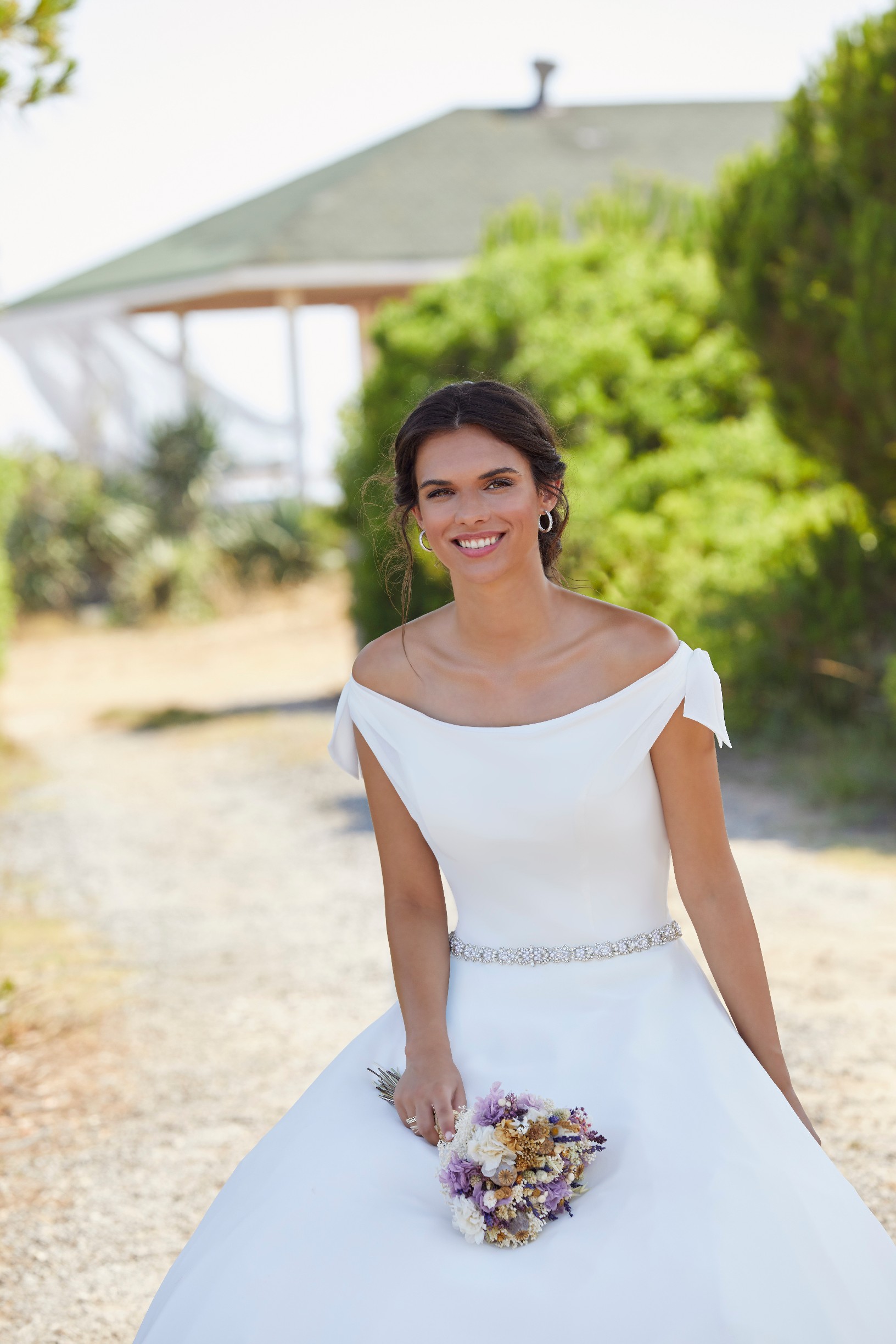 Brunette woman standing in garden in warm and sunny climate wearing off the shoulder ballgown wedding dress with delicate bow detail