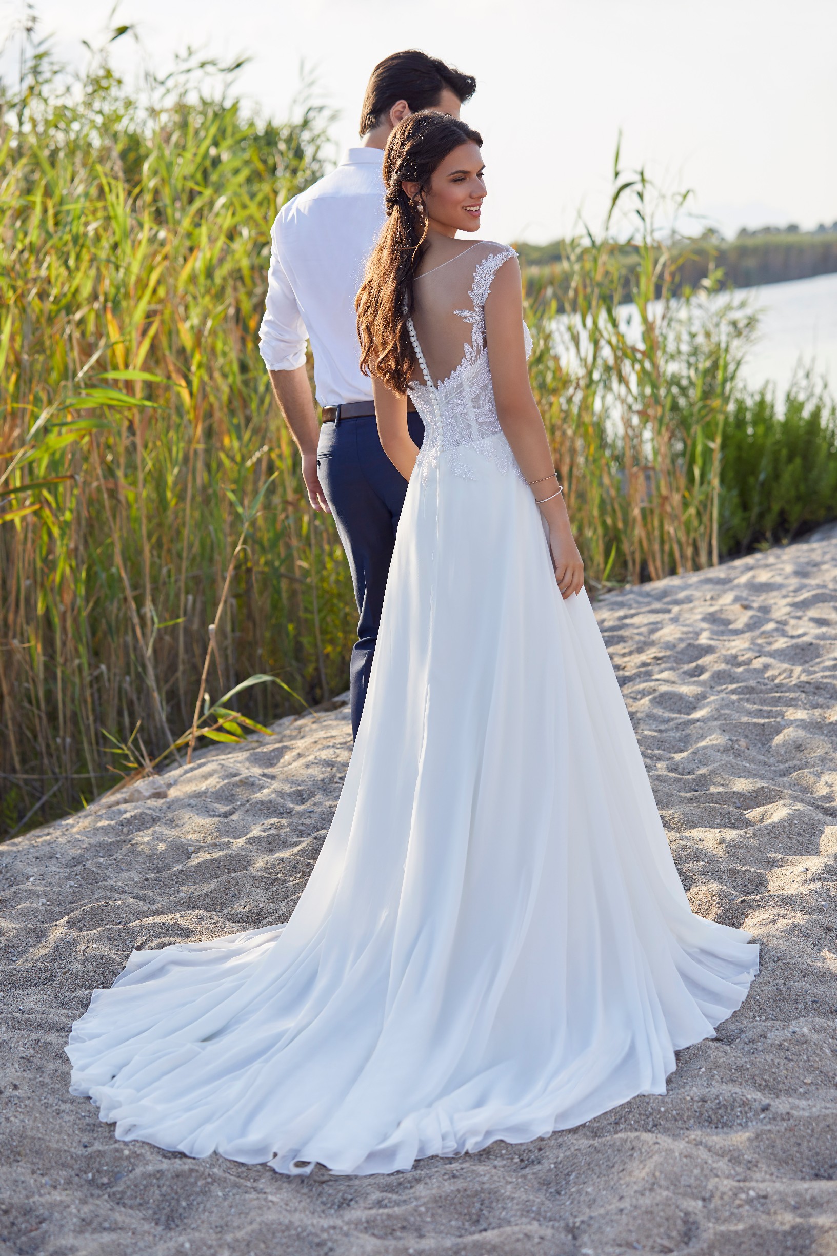 The back of a bride and groom stood by beach reeds. Bride wears Ronald Joyce 18413, a white A-line wedding dress with an illusion buttoned back, plain skirt and beaded tulle bodice