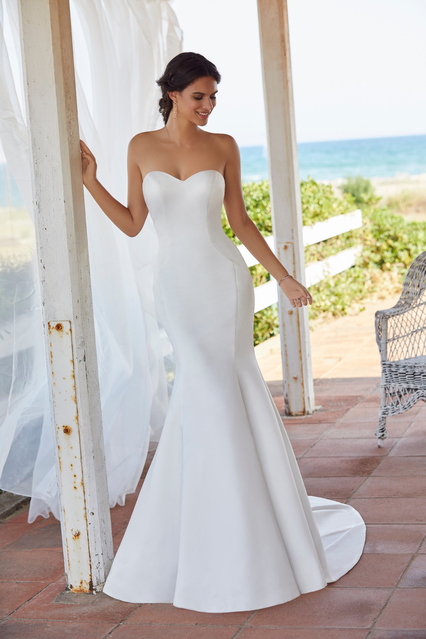 Brunette woman stands on porch beside the sea wearing simple and classic strapless wedding dress with fishtail skirt