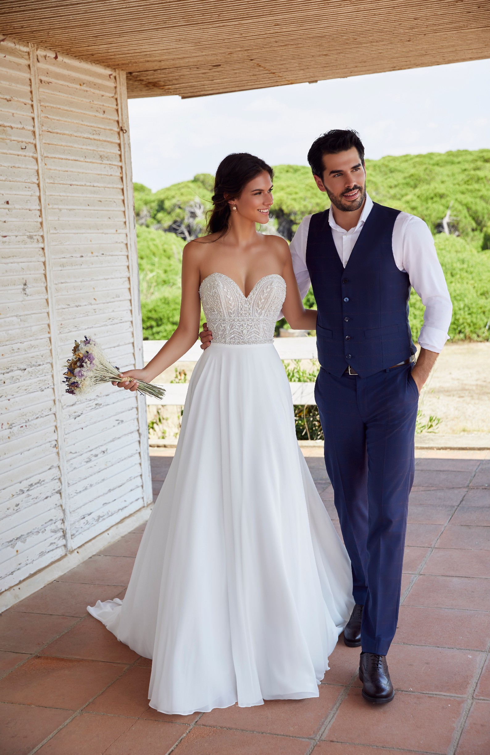 Bride and groom models stood on a terrace near greenery. Bride wears Ronald Joyce 18419, a strapless destination wedding dress with a beaded sweetheart bodice and plain skirt 