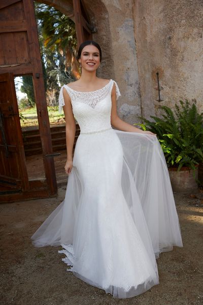 Model stood by a gate in Ronald Joyce 18455, a plain fit and flare wedding dress with lace tie shoulders, a sweetheart lace neckline and lace hem. Model also wears a sheer tulle A-line overskirt.