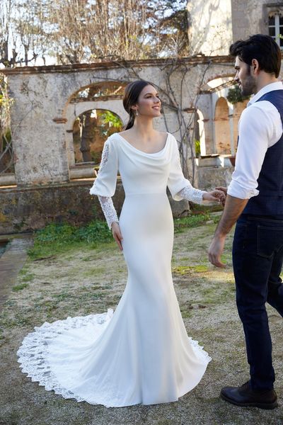 Model stood outside in Ronald Joyce style 18456, a plain ivory fit and flare winter wedding dress with a cowl neckline and long blouson sleeves with a bold lace cuff