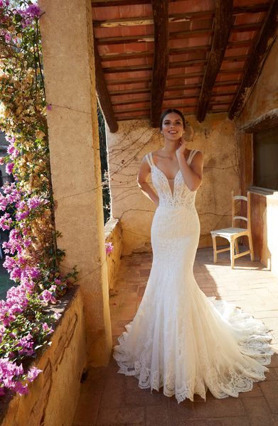 Model stood outside on a pink floral terrace in Ronald Joyce style 18467, one of our lace fit and flare wedding gowns with a plunging illusion neckline and double straps