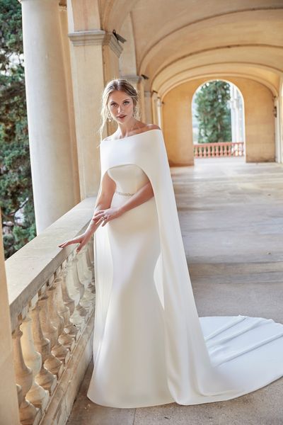 Model stood on a posh terrace in Ronald Joyce style 18501, a plain fit and flare wedding dress with a strapless sweetheart neckline and sparkle belt. Model also wears a matching cape