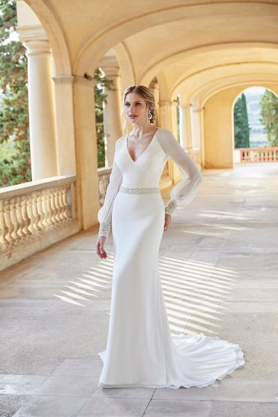 Model stood on a stately home terrace in Ronald Joyce 18506, a classic chiffon fit and flare wedding dress with v-neckline, long sheer sleeves and sparkle belt and cuff detail