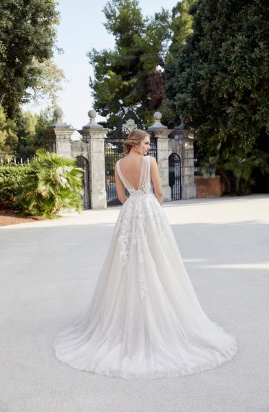 Back of model stood by black iron gates in Ronald Joyce style 18507, a vintage inspired ballgown wedding dress with lace embroidery, ruched tulle straps and an open v-shaped back