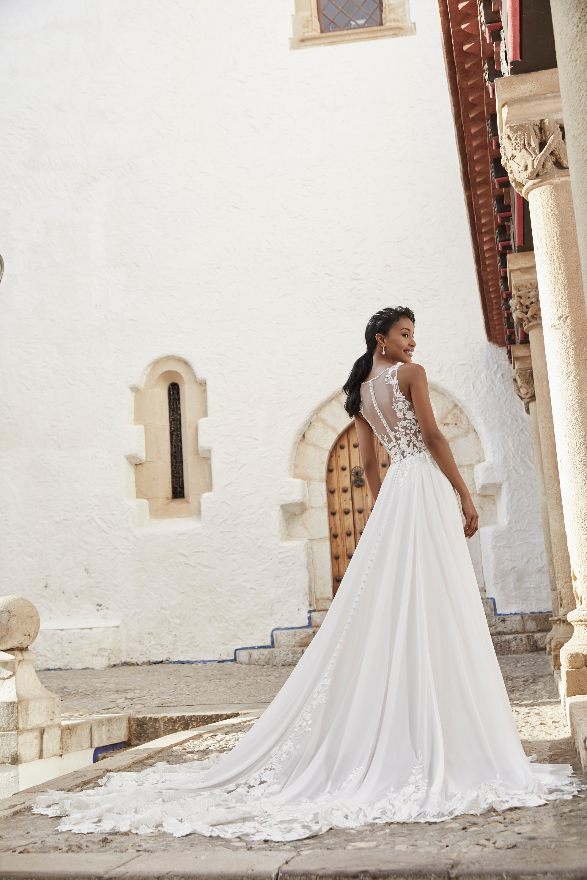 Woman stood outside historic villa wearing a-line wedding dress with lace embroidered train and illusion back with button details
