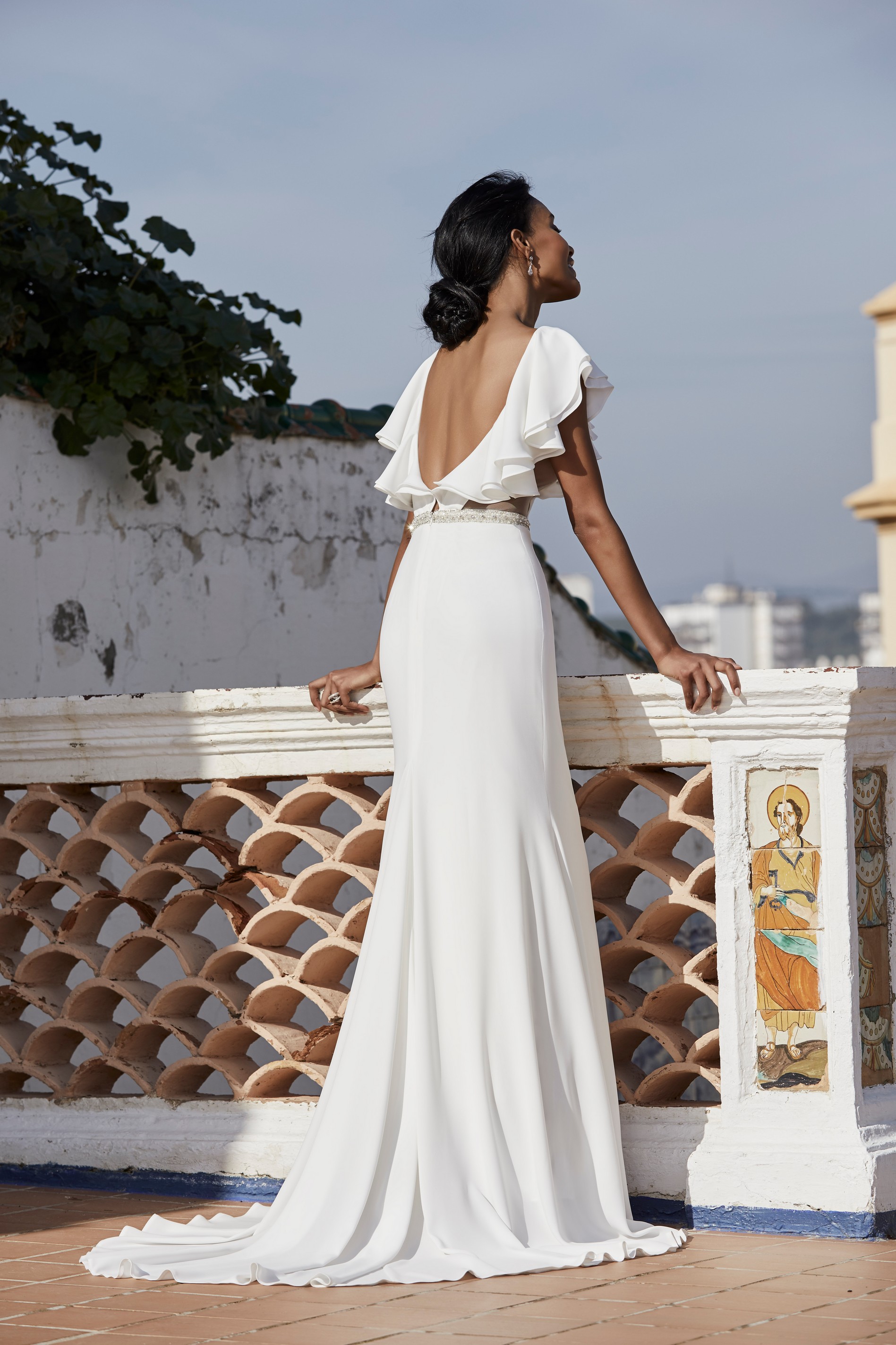 Rear profile of a black model on a Spanish terrace in Victoria Jane style 18553, a plain destination wedding gown with an open back, ruffle cap sleeves and sparkle belt 