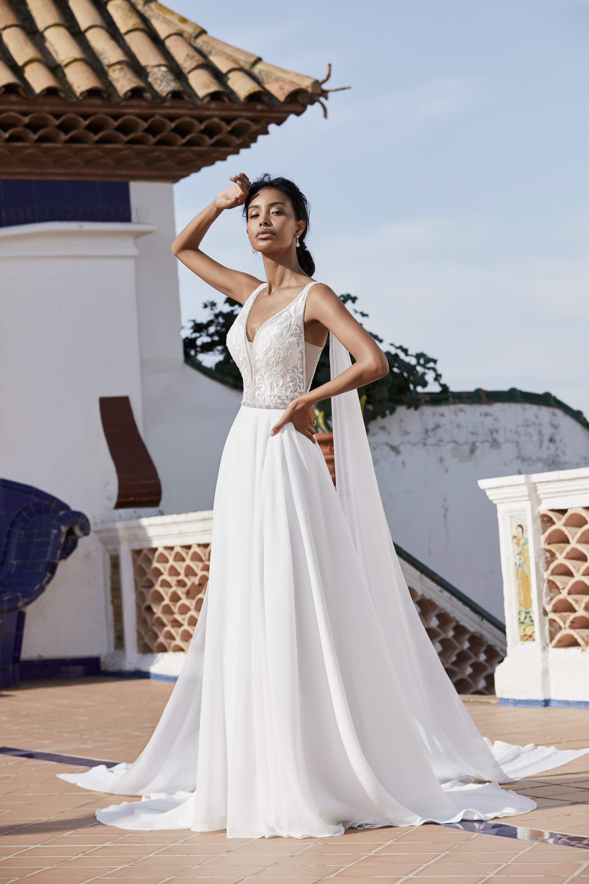 Model stood on a Spanish terrace in Victoria Jane 18555, an A-line destination wedding gown with a plunging neckline, lace body, plain floaty skirt and shoulder capes