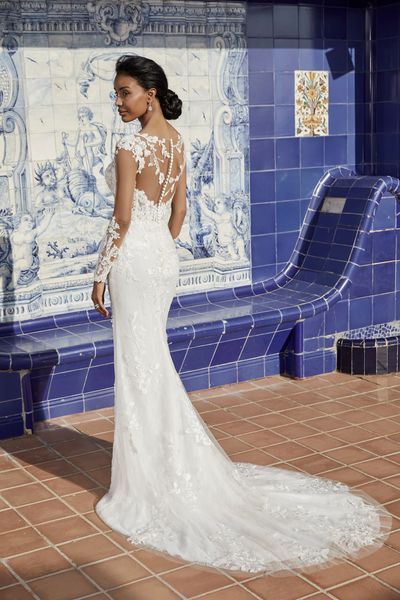 Back of model stood in front of a Spanish tiled wall in Victoria Jane 18556, a lace fit and flare destination wedding gown with a stunning sheer necklace back, and long lace sleeves