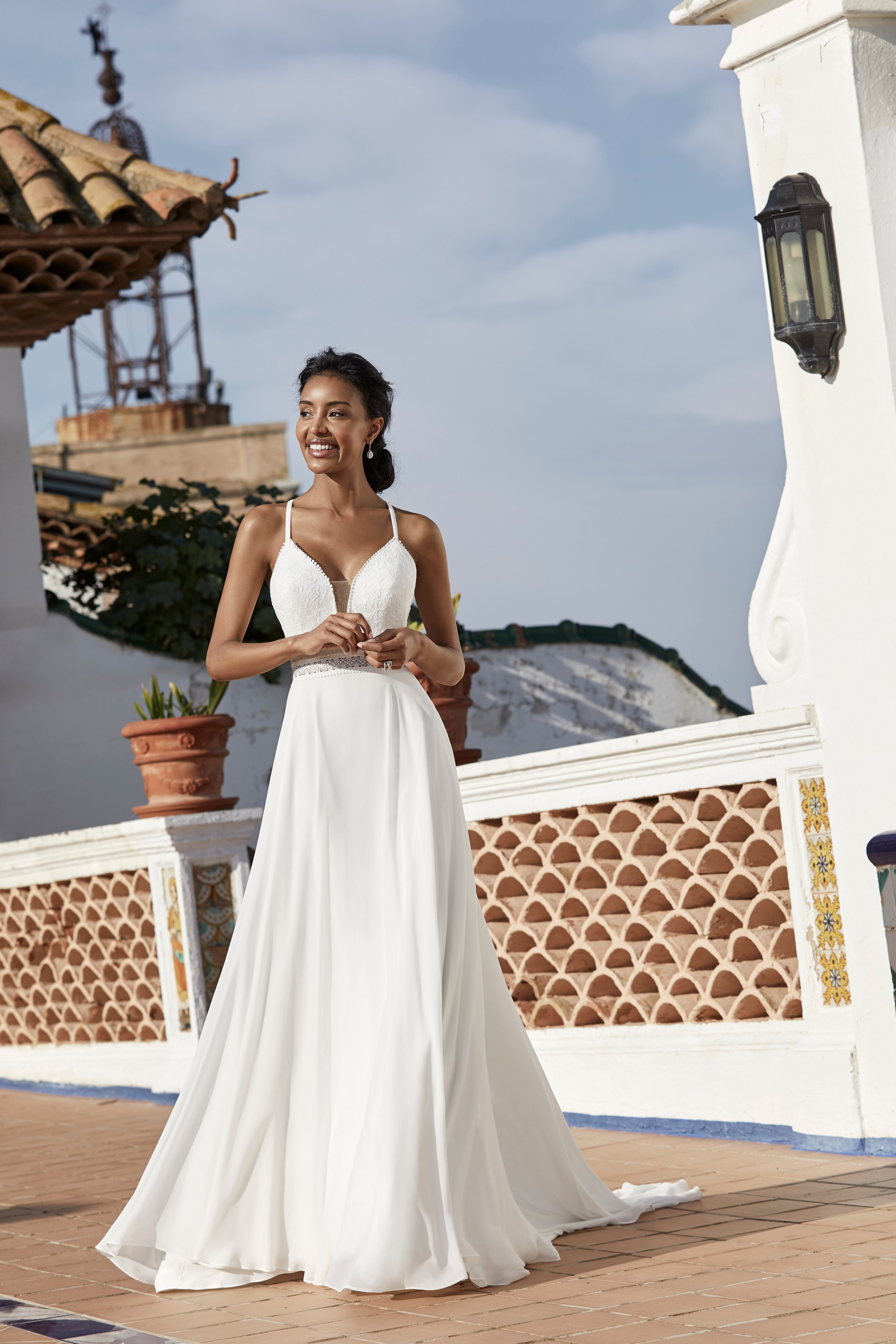 Model stood on a Spanish terrace in Victoria Jane style 18560, a plain A-line destination wedding gown with a plunging illusion neckline, delicate straps and crochet waistband 