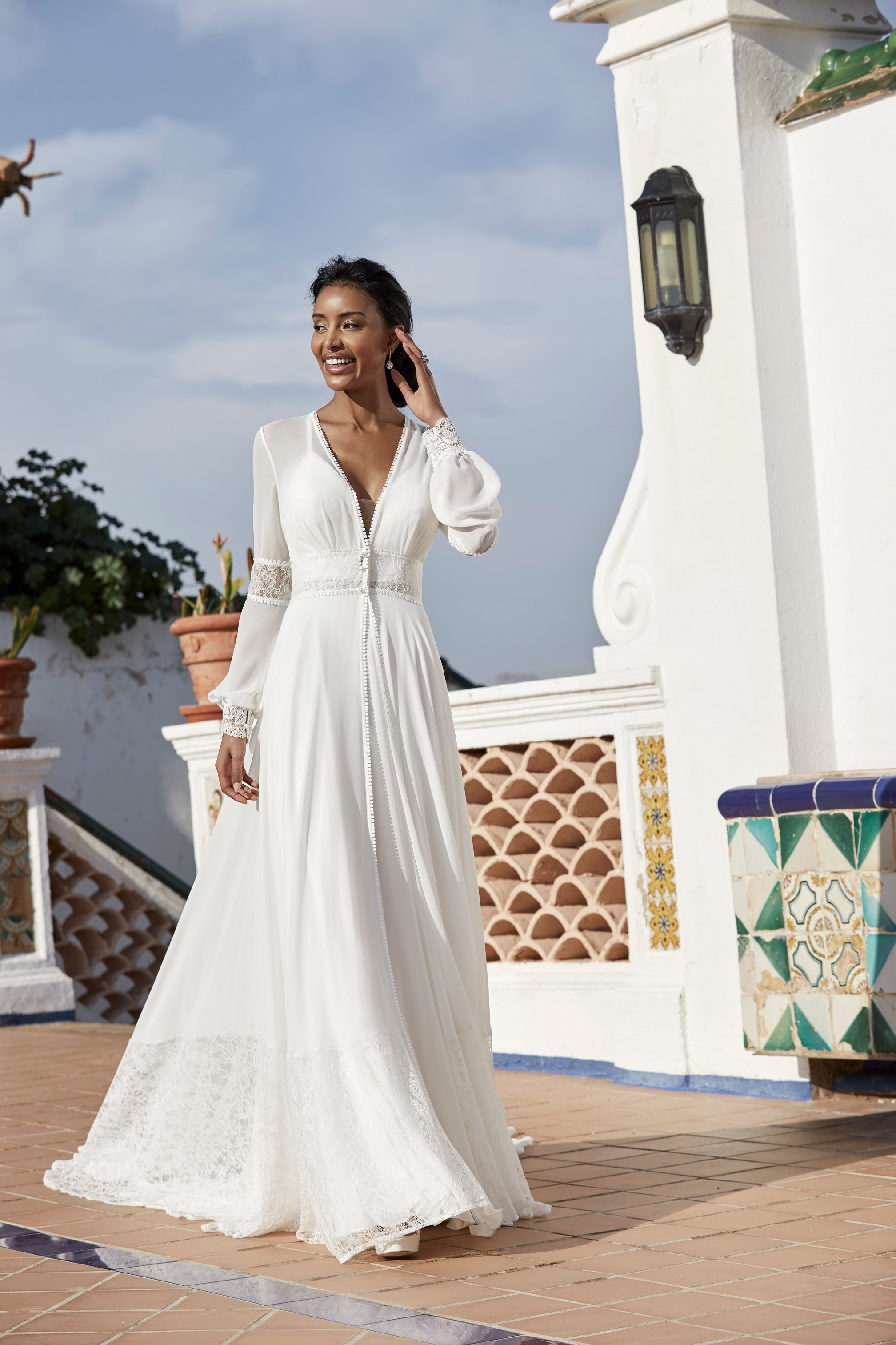 Model stood on a Spanish terrace in Victoria Jane style 18560, a plain A-line destination wedding gown and matching long sleeve chiffon cape as style 18561