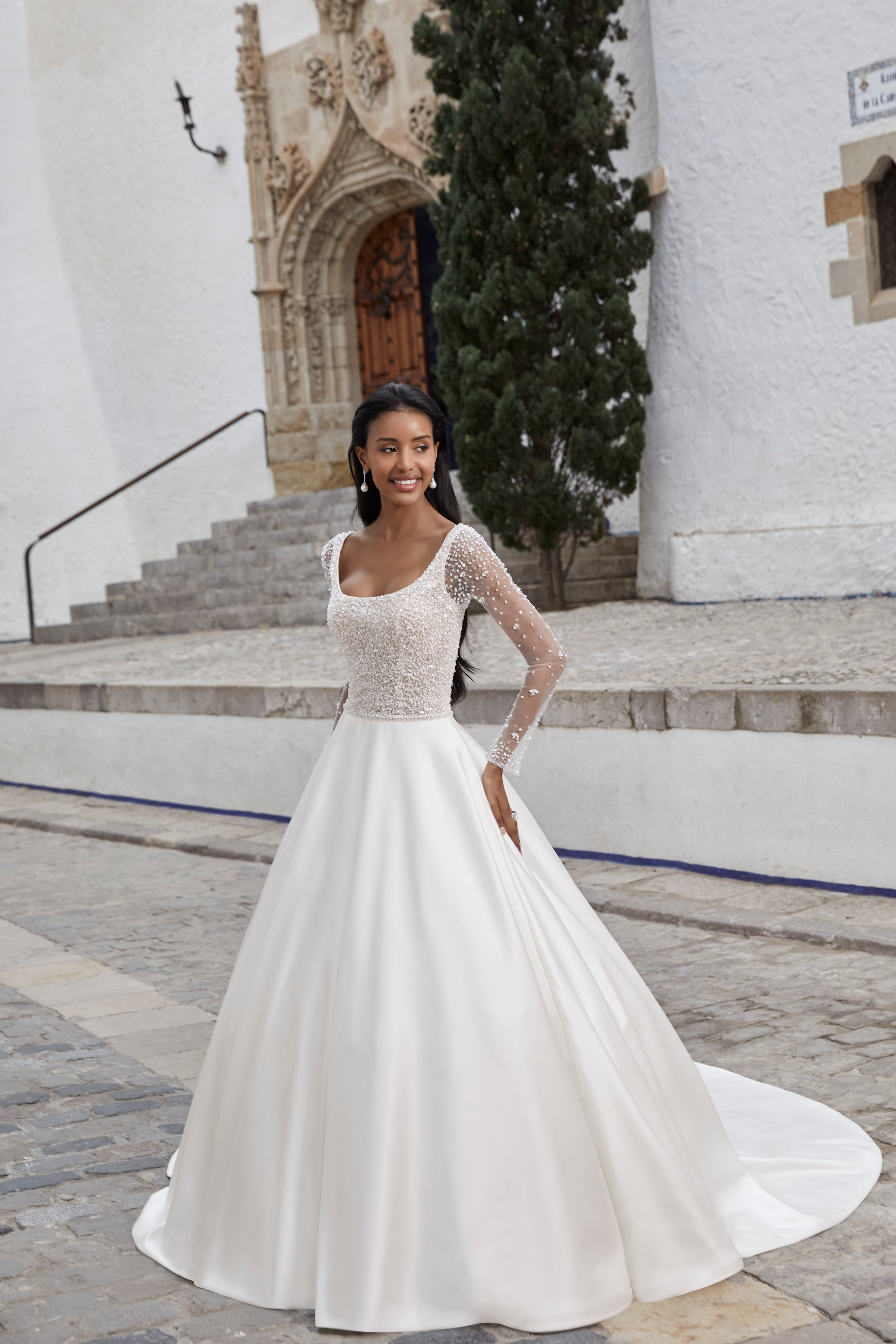 Woman standing in front of scenic church and tree wearing princess ball gown wedding dress with beaded bodice and long sleeves