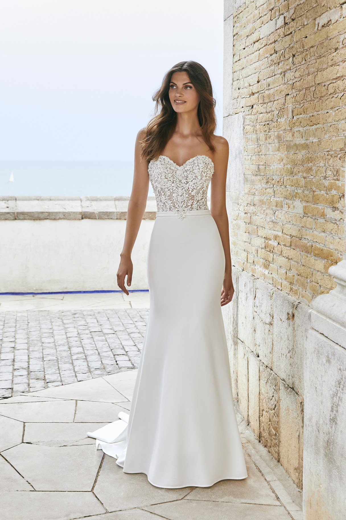 Model stood by a stone wall in Ronald Joyce style 18604, an elegant fit and flare wedding dress with a beaded sweetheart bodice, plain skirt and matching delicate belt