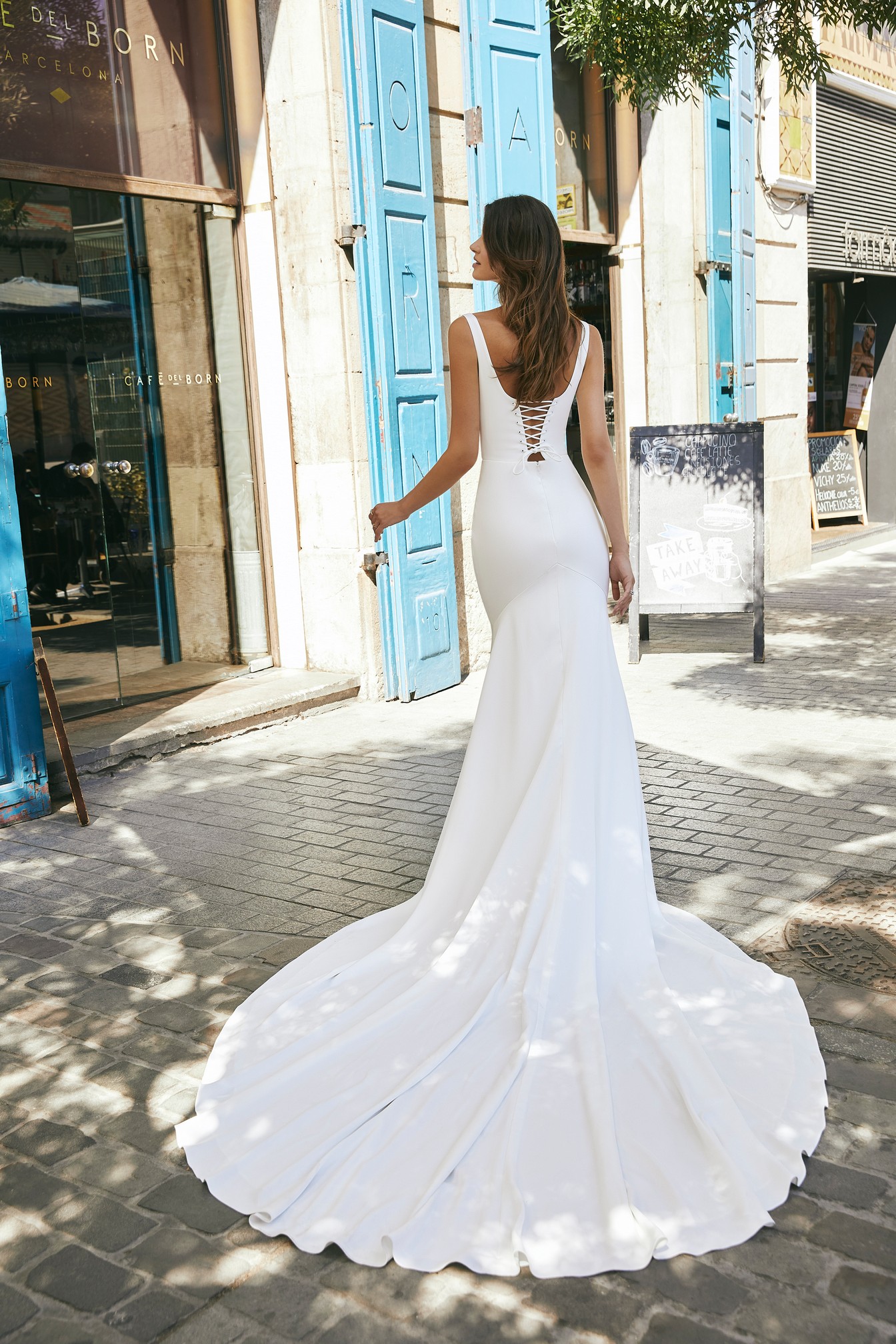 Back image of woman standing in front of Barcelona cafe bar in crepe wedding dress with long train