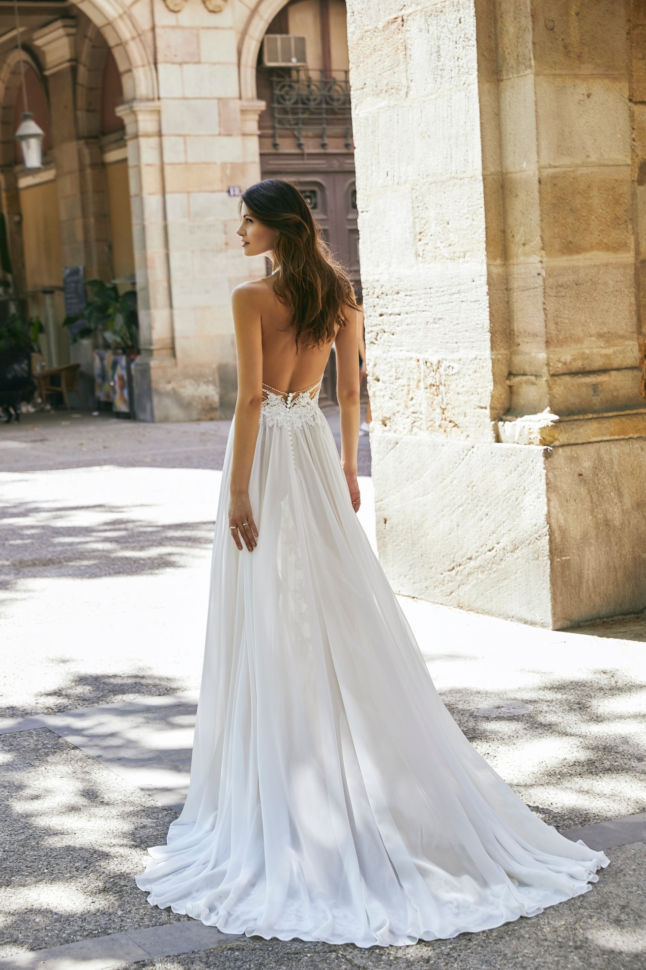 Back profile of model in Ronald Joyce destination wedding dress style 18611, a boho lace illusion halter neck dress with a plain chiffon skirt and open back