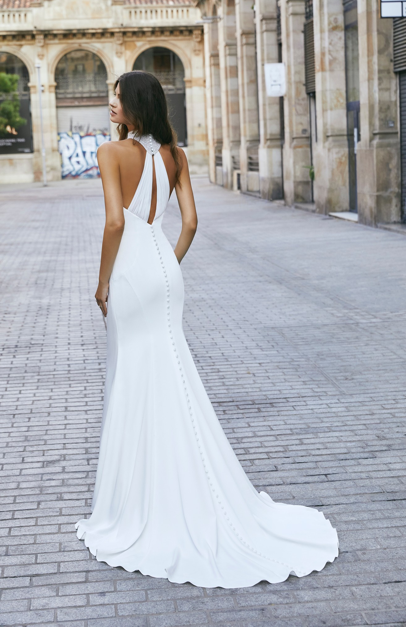 Lady stands in Barcelona plaza wearing classic crepe fit and flare wedding dress with unique back detail