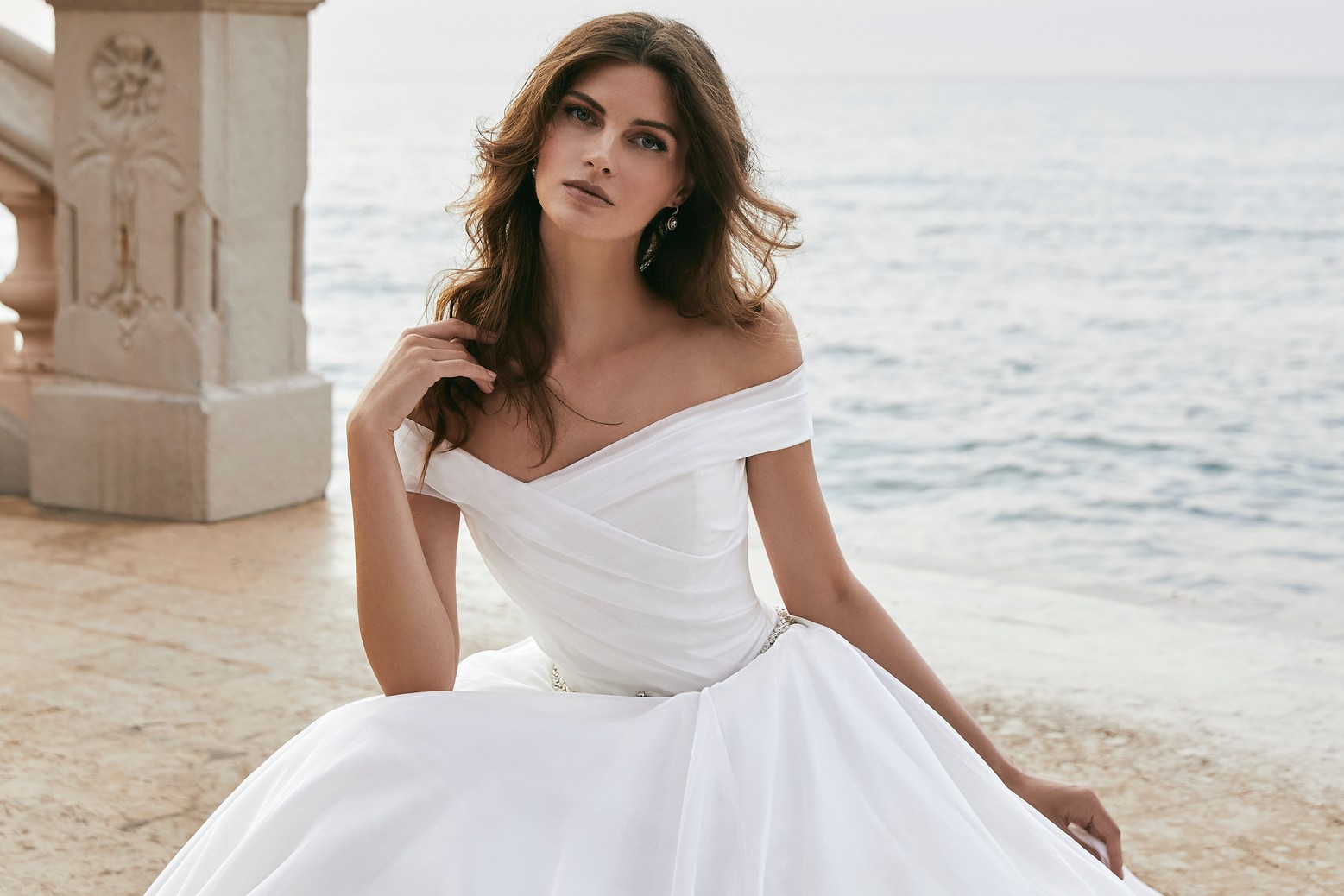 Close up of a brunette model sat on a stone floor by the sea in Victoria Jane 18619, a plain ivory ballgown wedding dress with a bateau off-the-shoulder neckline and a delicate sparkly diamante belt