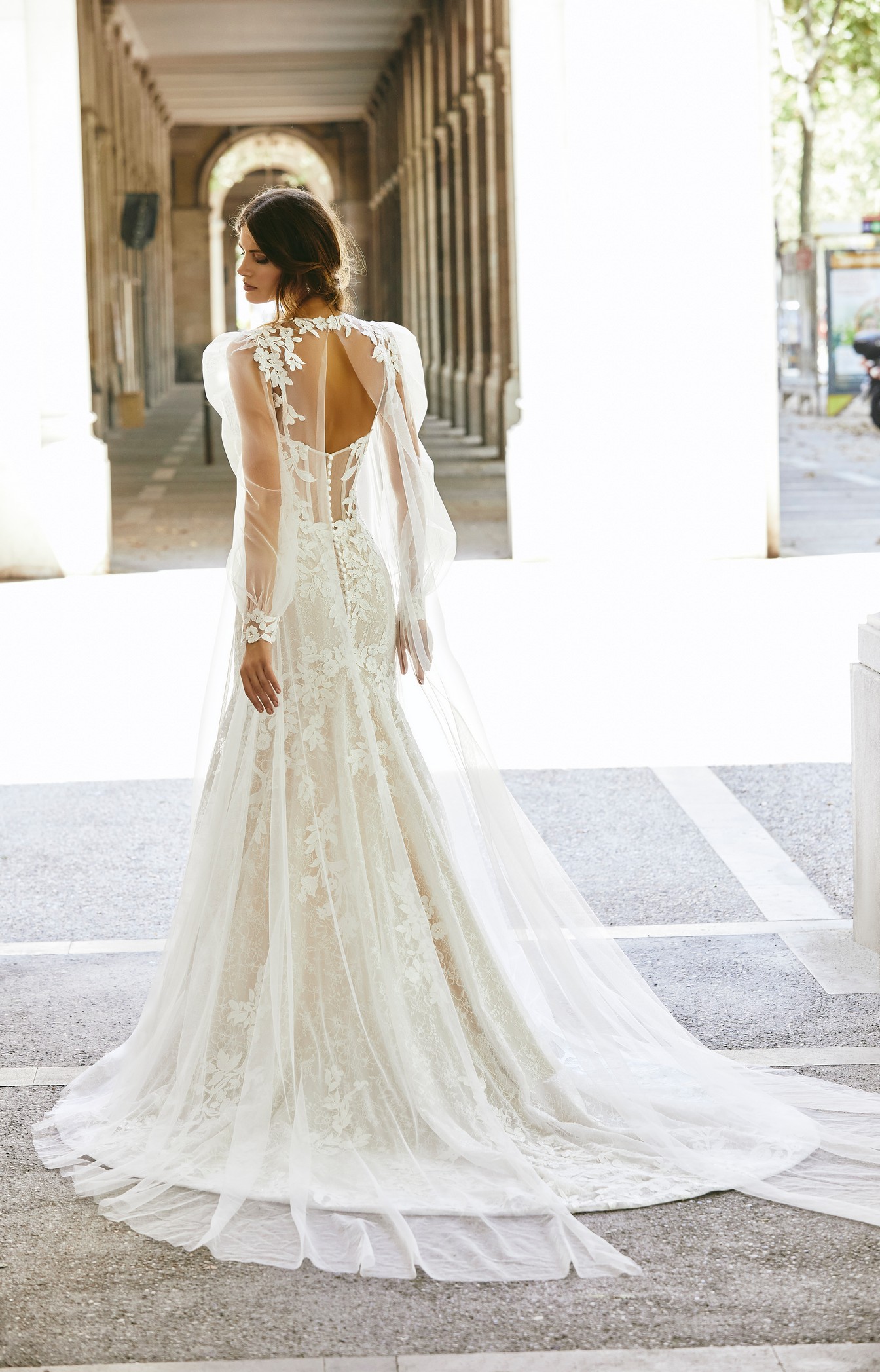 Woman standing between pillars wearing fit and flare lace wedding dress with illusion back and tulle coat 