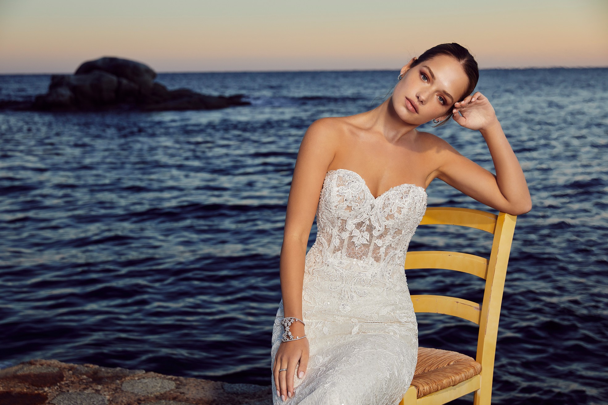 Woman sat on wooden chair next to the sea wearing strapless lace wedding gown with sweetheart neckline